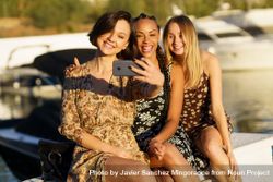 Three smiling women sitting on wharf taking selfie 5Xy2o4