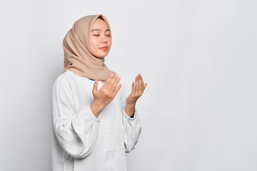 Muslim woman in headscarf and light blouse with eyes closed and hands up in prayer with copy space