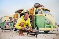 Tanned man cooking on camping stove beside parked mini van