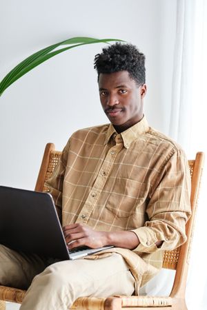 Black man with laptop resting on chair on white background