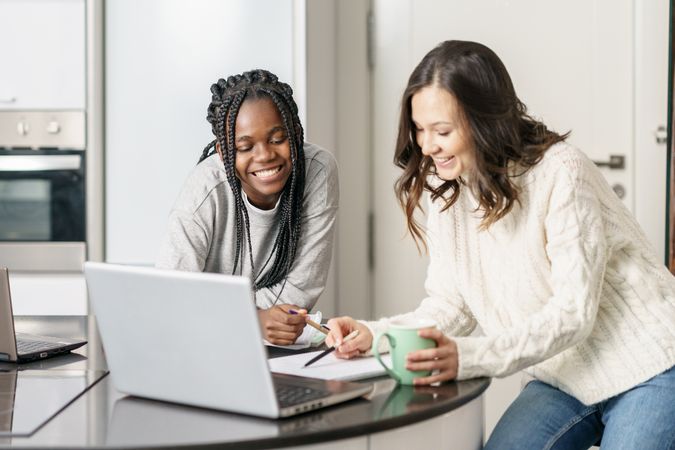 Two women smiling and working together at the kitchen table on a laptop