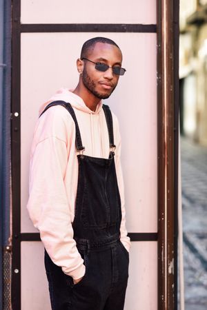 Male in denim overalls and earrings with hands in pockets against a pink background