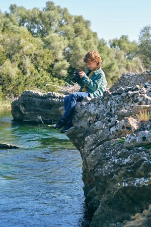 Boy taking photo while sitting on rocky cliff near water