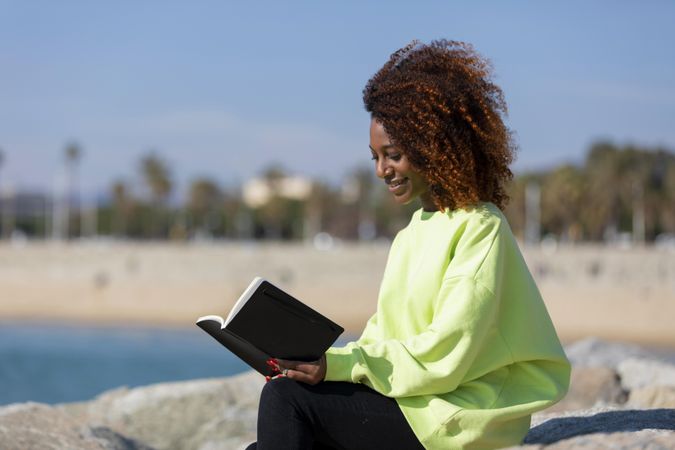 Female in bright green shirt sitting on the coast on sunny day with a book