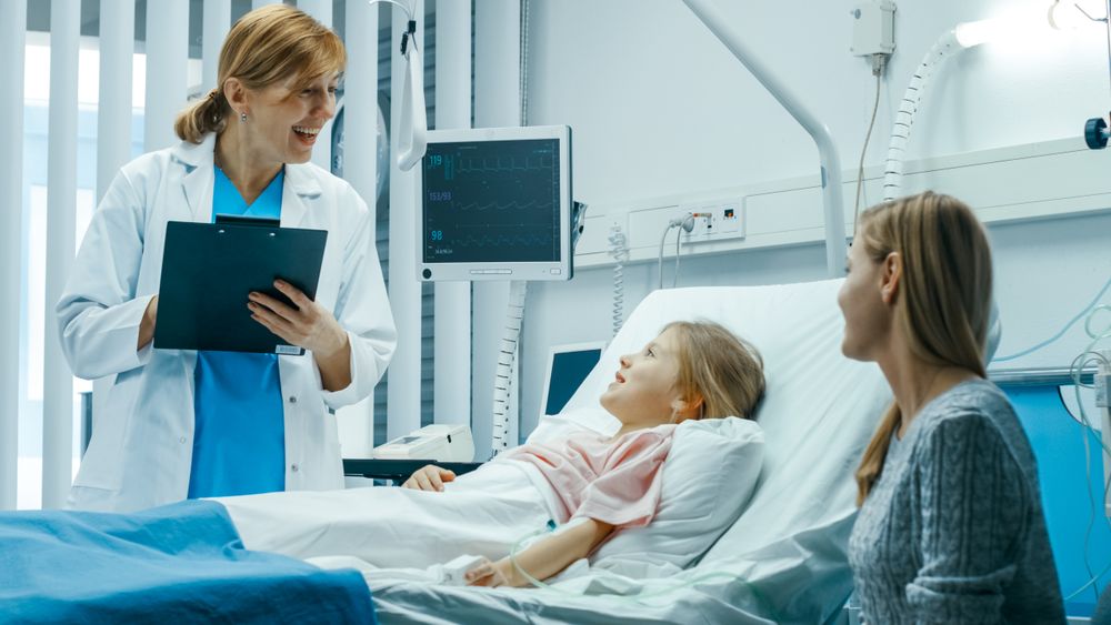 Recovering Little Girl Lies on a Bed In the Hospital, Talks to a Friendly Doctor With Clipboard, Mother Sits Beside