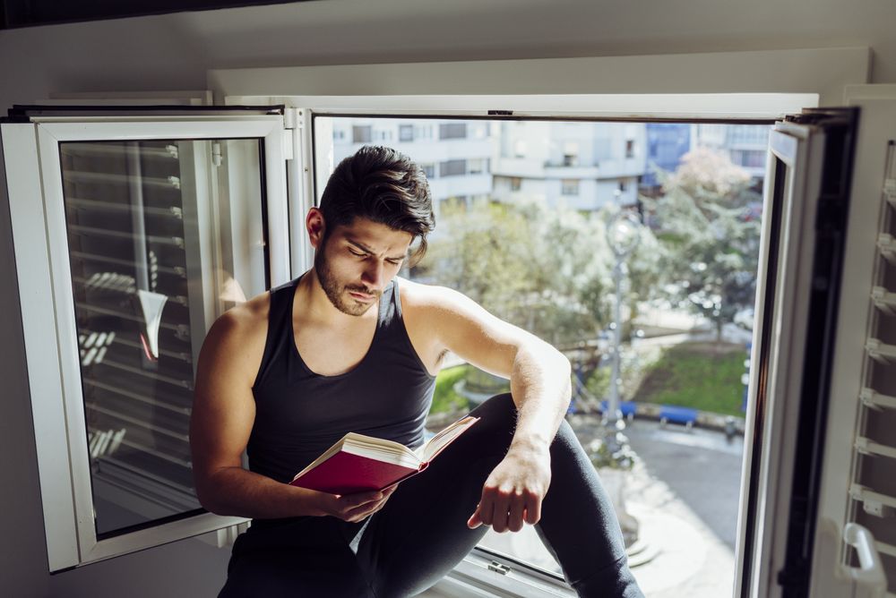 Young male sitting on window and reading book