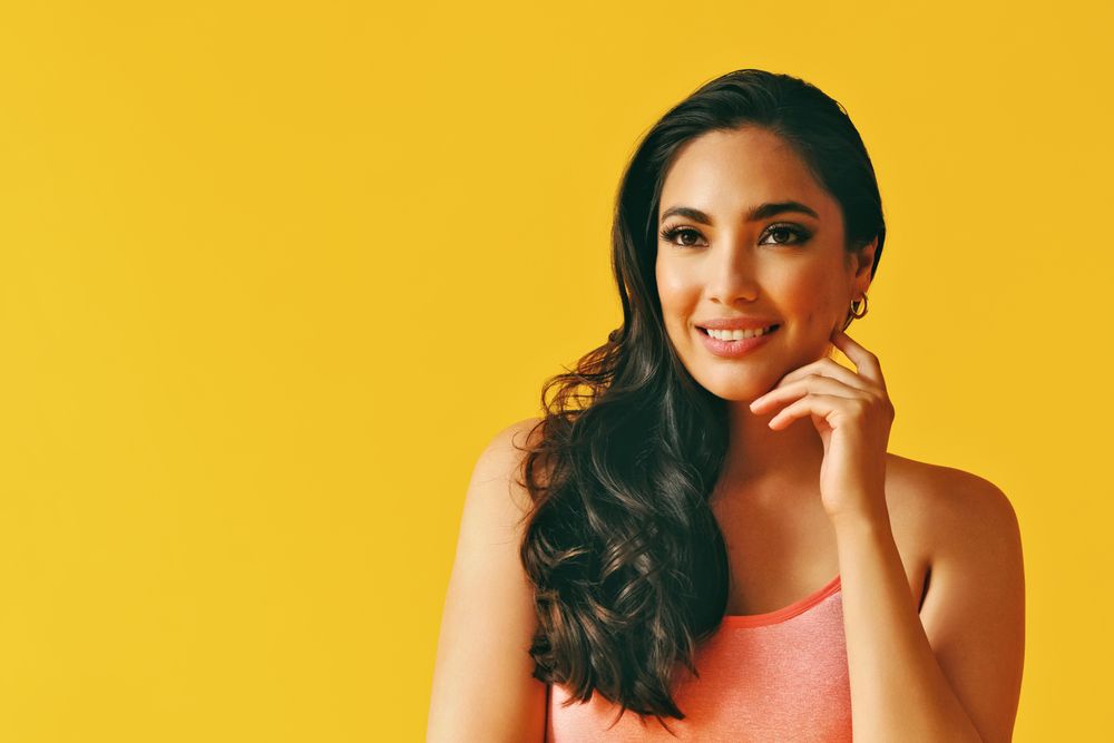 Headshot of smiling Hispanic woman looking away in yellow room, with hand up to her cheek