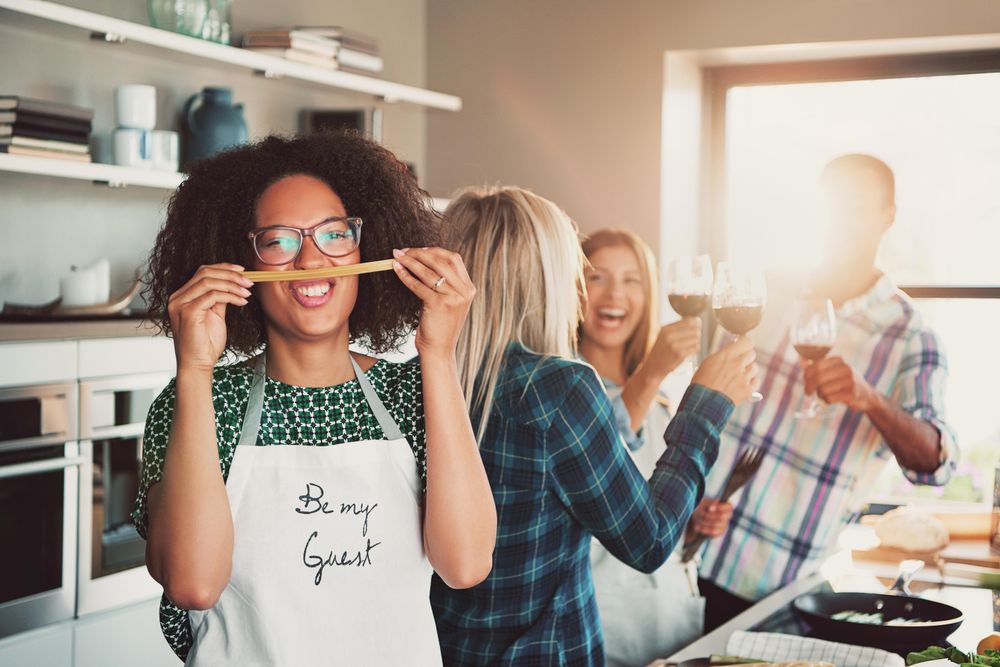 Woman makes a silly face with pasta while her friends toast with red wine behind