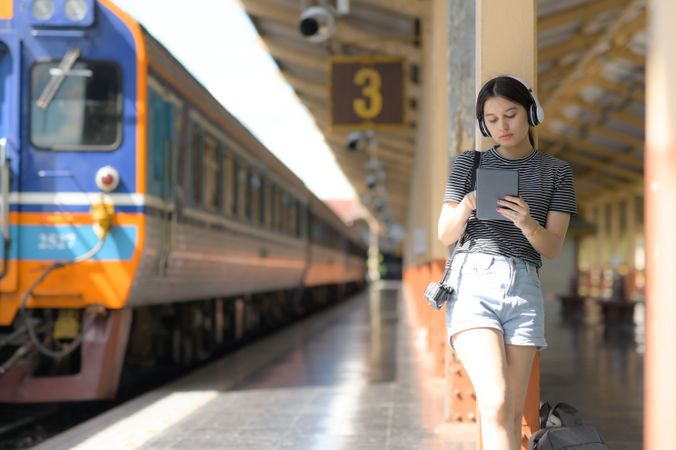 A woman wearing headphones listening to music from an app on a tablet while waiting for train