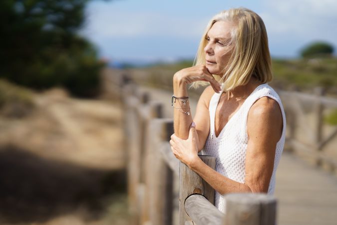 Calm older female with grey hair with hand on her chin on wooden walkway near the coast