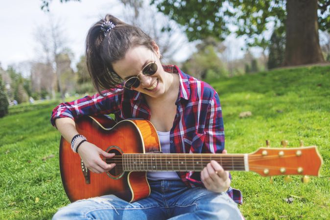 Front view of a young hipster woman wearing sunglasses, sitting on grass in a park while enjoying playing guitar