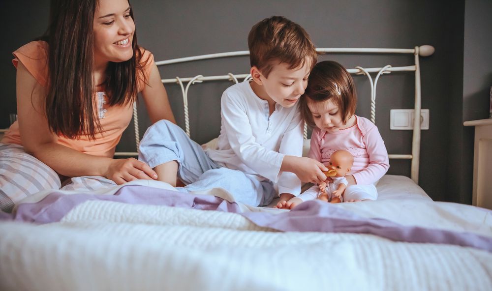 Portrait of happy children feeding doll with a cookie sitting in bed with their mother