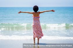 Rear shot of female in maxi dress with outstretched arms facing blue ocean 0LmyD5