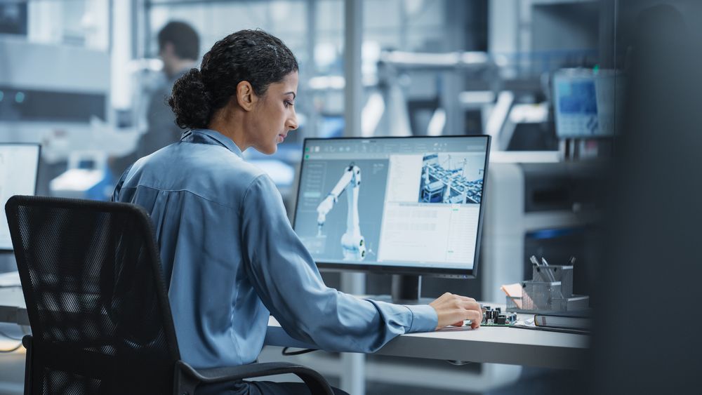 Hispanic Female Automation Engineer Using Desktop Computer to Control Automated Production With Robotic Arms On Electronics Factory, While Indian Assembly Line Worker Using Solder Jet Printer.