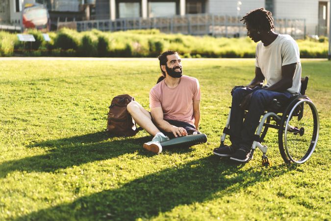 Two adults with disabilities, one with prosthetic leg, the other sitting in a wheelchair, relaxing carefree chatting on the grass on a summer day