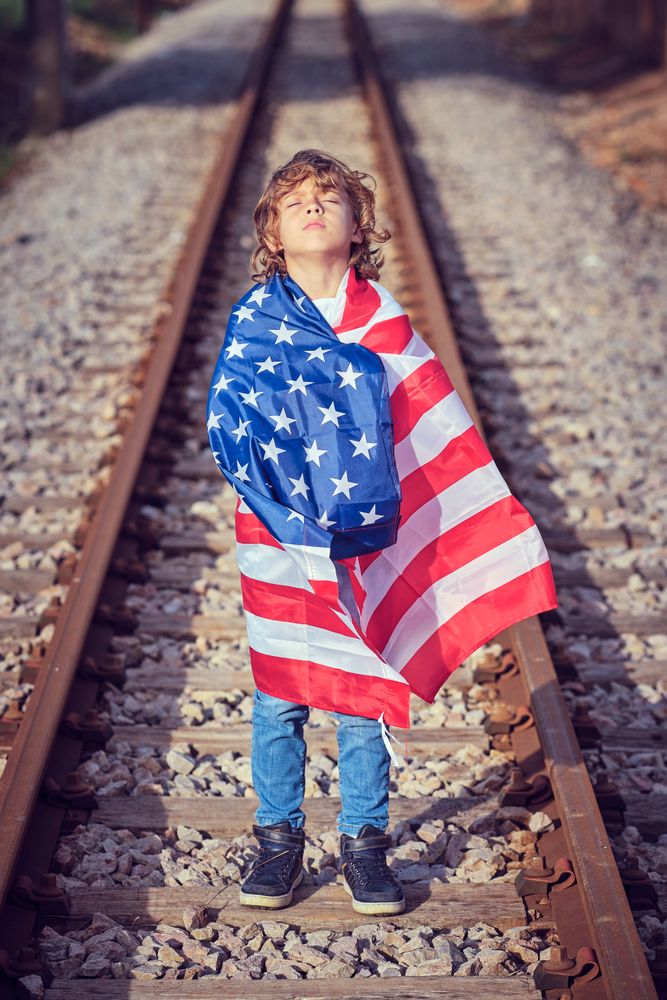 Adorable preteen boy wrapped in American flag standing on railwa