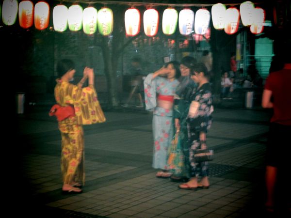 Three women in kimonos standing under lit lanterns at night