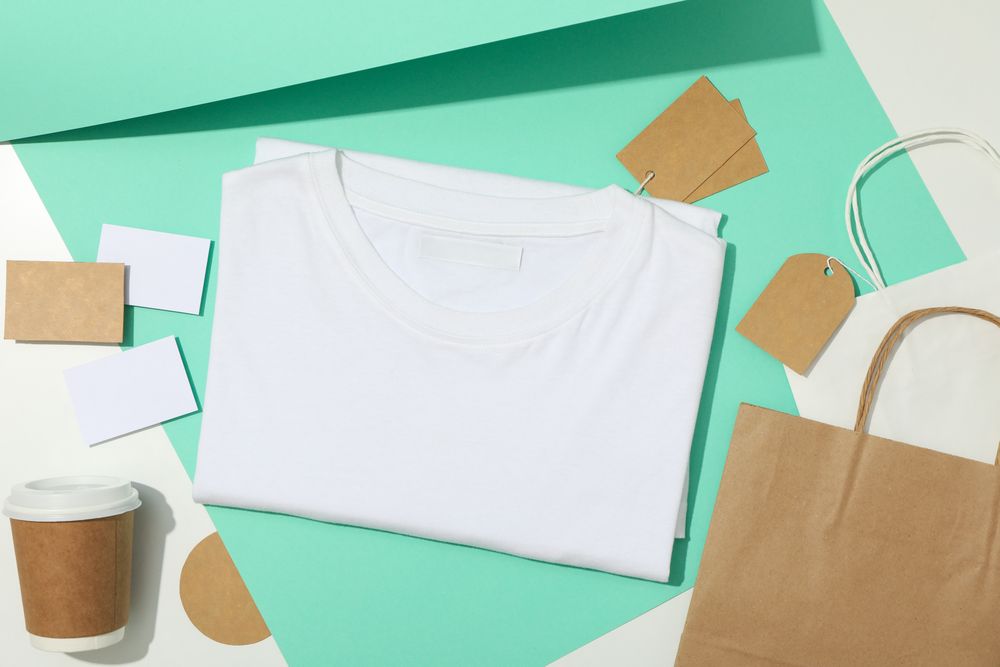 Blank t-shirt, tags, paper cup and paper bags on mint and light background, space for text