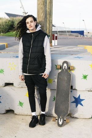 Teenage girl leaning against a wall next to her longboard while looking at camera