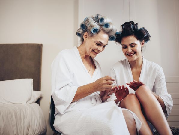Two women in bathrobe with hair rollers sitting together at home doing their nails