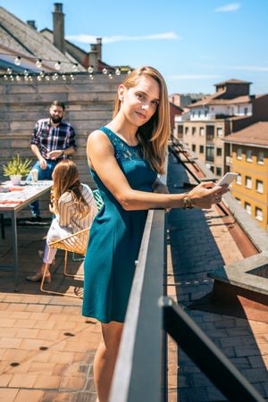 Young woman with green dress looking at camera while holding mobile on rooftop