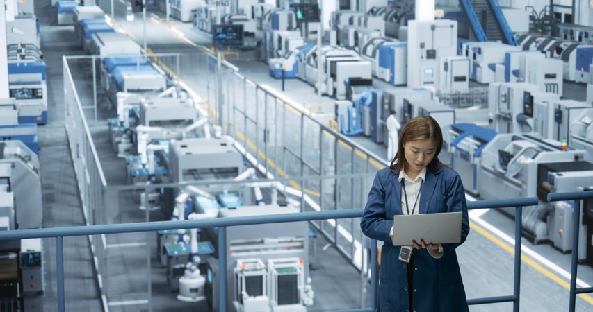 Japanese Factory Specialist Working on Laptop at a Futuristic Industrial Manufacture with Autonomous AI Robots. Project Manager Monitoring Data on a Computer, Checking Reports and Planning Research