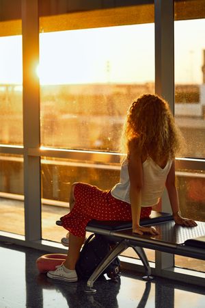 Stylish cheerful woman sitting on metal bench in glass hallway of airport in Texas
