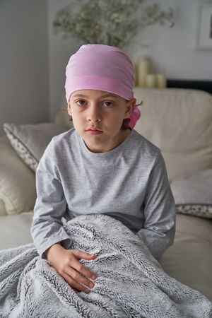 Peaceful sick little boy in bandana sitting on bed and looking a