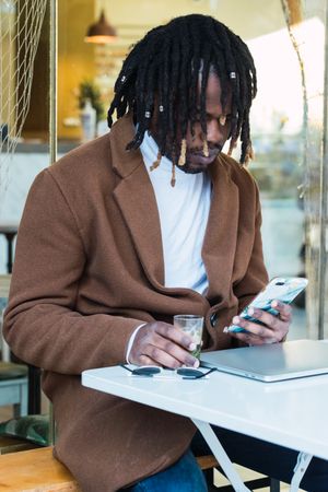 Black man with laptop browsing on mobile phone on restaurant terrace
