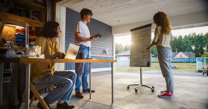 Hispanic Female CEO Using Whiteboard To Explain Business Objectives To Two White Male Colleagues With Laptop And Tablet.