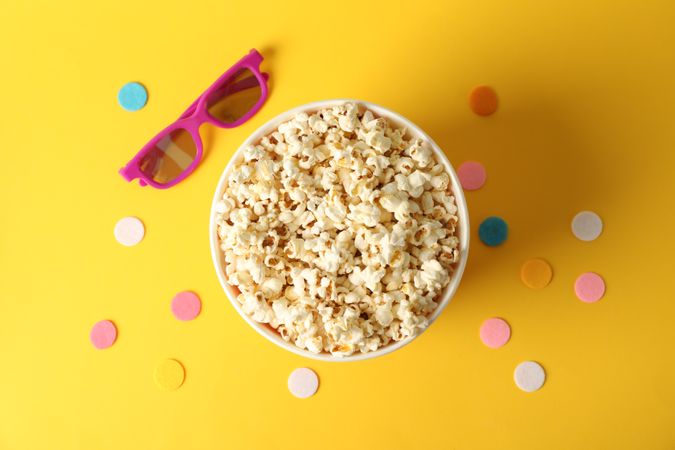 Striped bucket with popcorn and 3d glasses on yellow background, top view