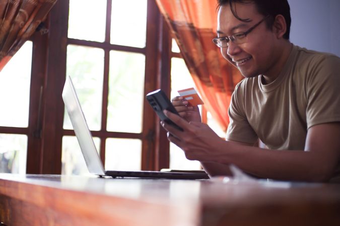 Asian Male with shirt doing online transaction in living room with credit card smartphone and laptop