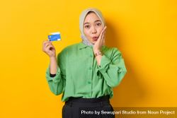 Muslim woman in headscarf and green blouse holding credit card and with hand to cheek in surprise bEaL7b