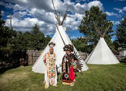 Two Native American men in full traditional regalia standing outside with teepees bE9V1b