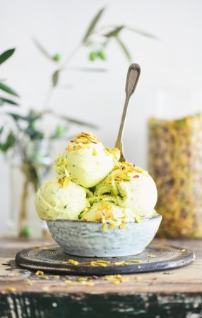 Bowl of melting pistachio ice cream with spoon, and light background with leaves and glass jar