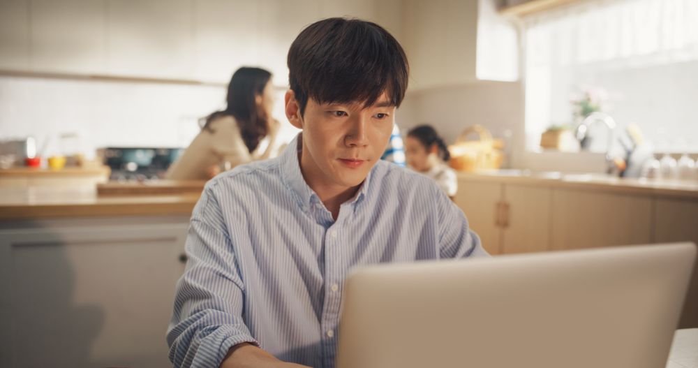 Portrait of Korean Family at Home: Young Father Using Laptop Computer for Working Remotely, With his Wife and Kids Preparing Lunch in the Background.