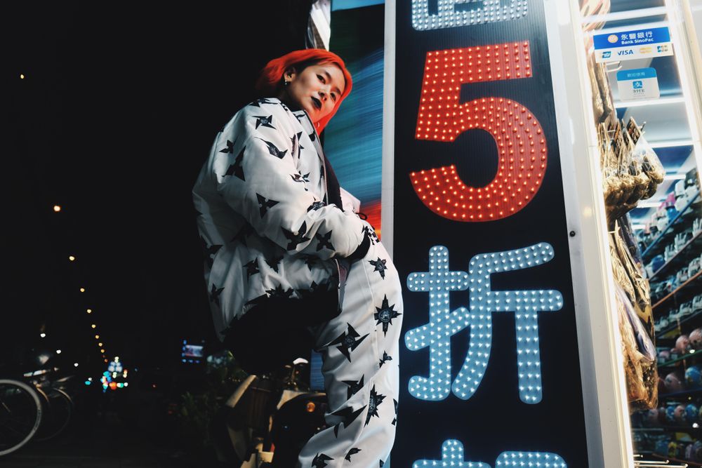 East Asian woman with red hair in casual outfit posing beside building at night