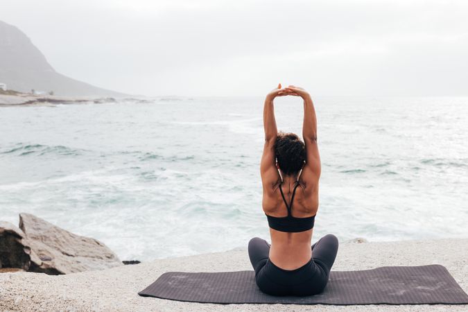 Female sitting next to the ocean sitting crossed legged stretching arms up