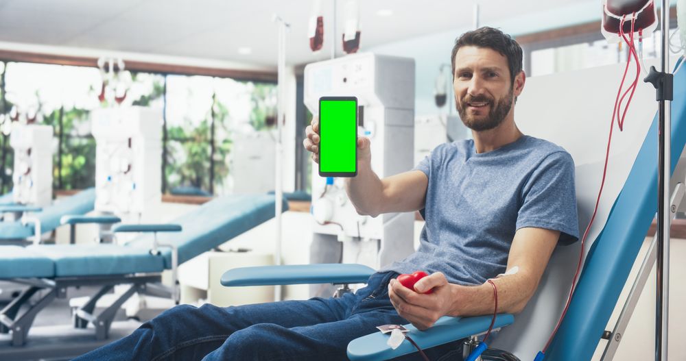 Man Sitting In The Chair, Squeezing Red Ball To Pump Blood Into Bag And Showing Green Screen Chromakey Smartphone To Camera.