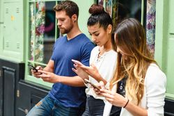 Two female and one male friend looking down at their phones by green wall