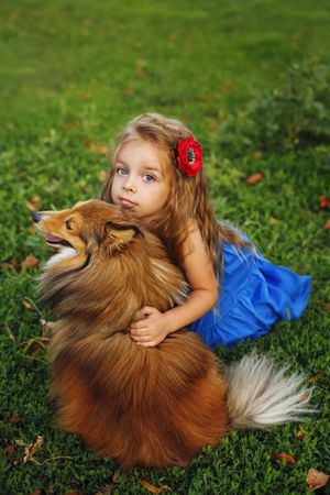 Happy child in blue dress hugging dog in the grass