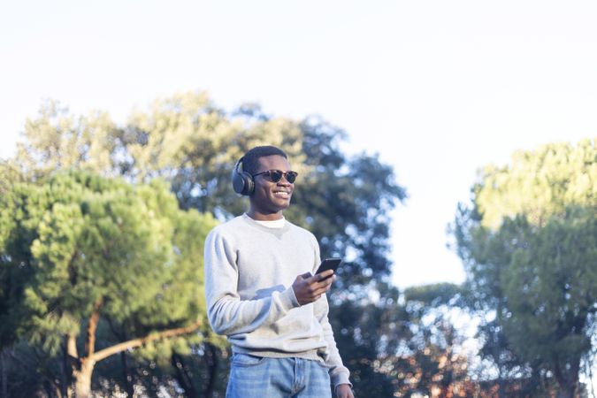 Happy Black male with sunglasses standing in park with phone