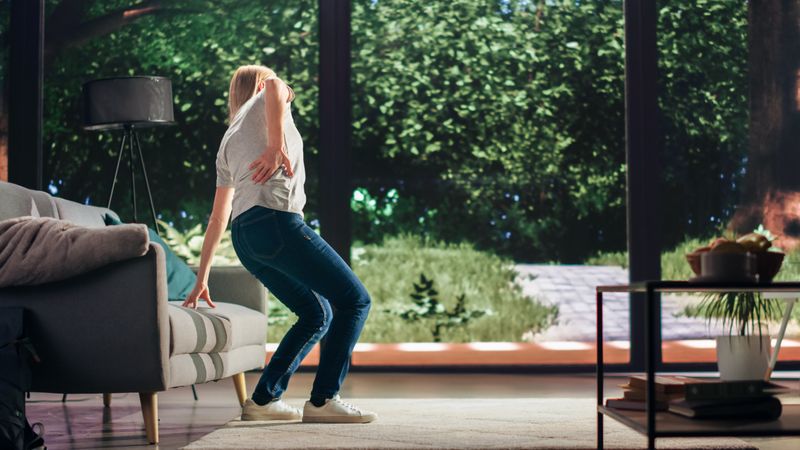 Young Adult Female Standing Up from a Couch at Home and Experiencing Back Pain in a Result of Spine Trauma.
