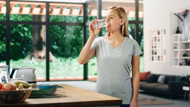 Young Adult Woman with Blond Hair Wearing Gray V-Neck T-Shirt, Drinking a Glass of Water After Lunch.