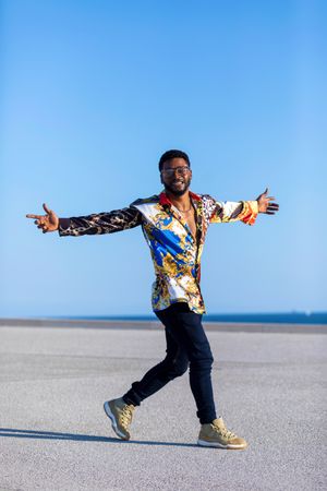 Black man wearing eyeglasses and loud shirt against blue sky walking with open arms