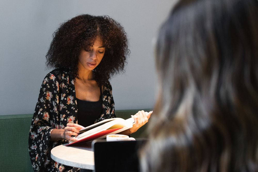 Black woman reading book against crop girlfriend in cafe