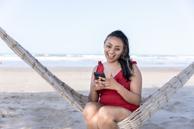 A young Hispanic woman is chatting with her phone on a hammock at the beach