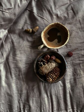 Top view of coffee in ceramic mug with bowl of pine cones on bed with grey sheets