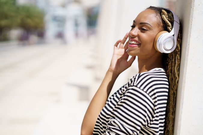Smiling female leaning on wall and listening to headphones on sunny day