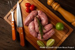 Top view of raw sausages arranged on wooden board with utensils and rosemary, lime and tomatoes 5QZGm4